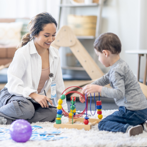 Therapist smiles at child playing with colorful bead maze on a carpeted floor.
