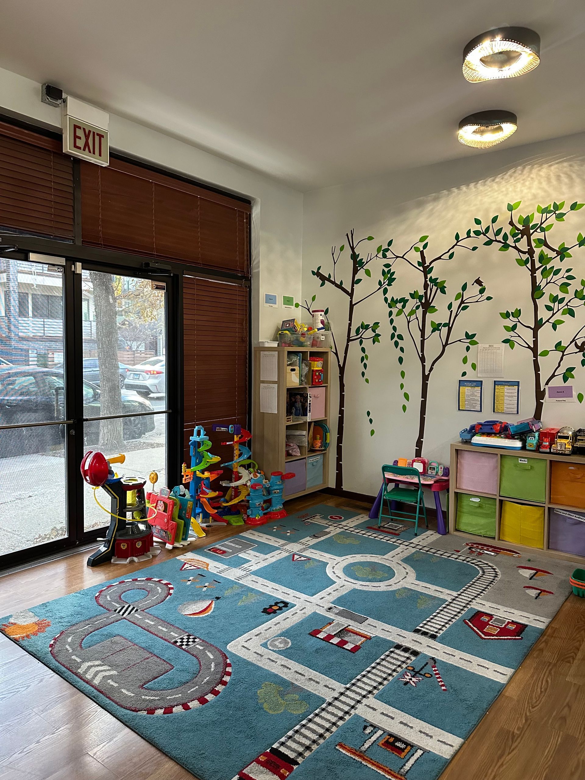 Playroom with road-themed rug, toys, bookshelves, and tree wall decals near an exit door.