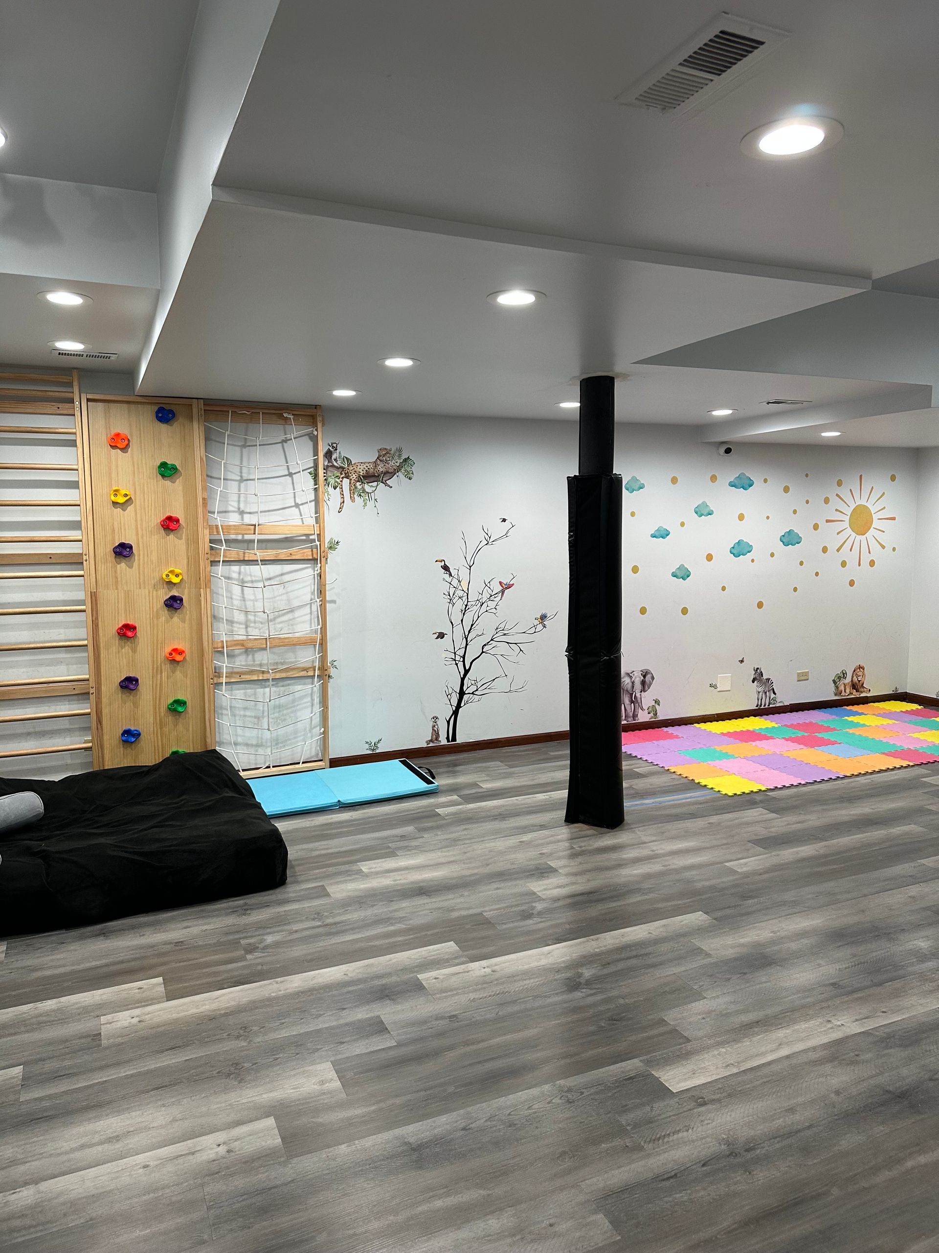 Basement play area with climbing wall, black support column, colorful foam tiles, wood-look flooring, and a sun and cloud decal.