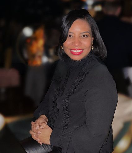 Woman in black top smiles, hands clasped. Red lipstick. Evening setting, slightly blurred background.