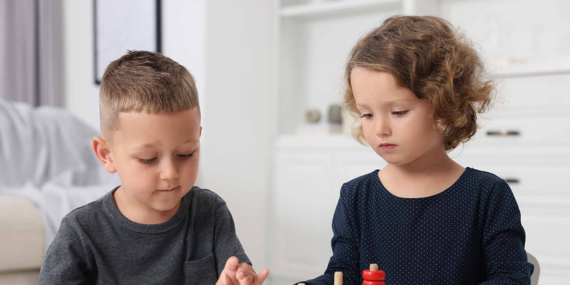 Two children sitting at a table looking down. One has short brown hair and a gray shirt. The other has curly brown hair and a blue shirt.