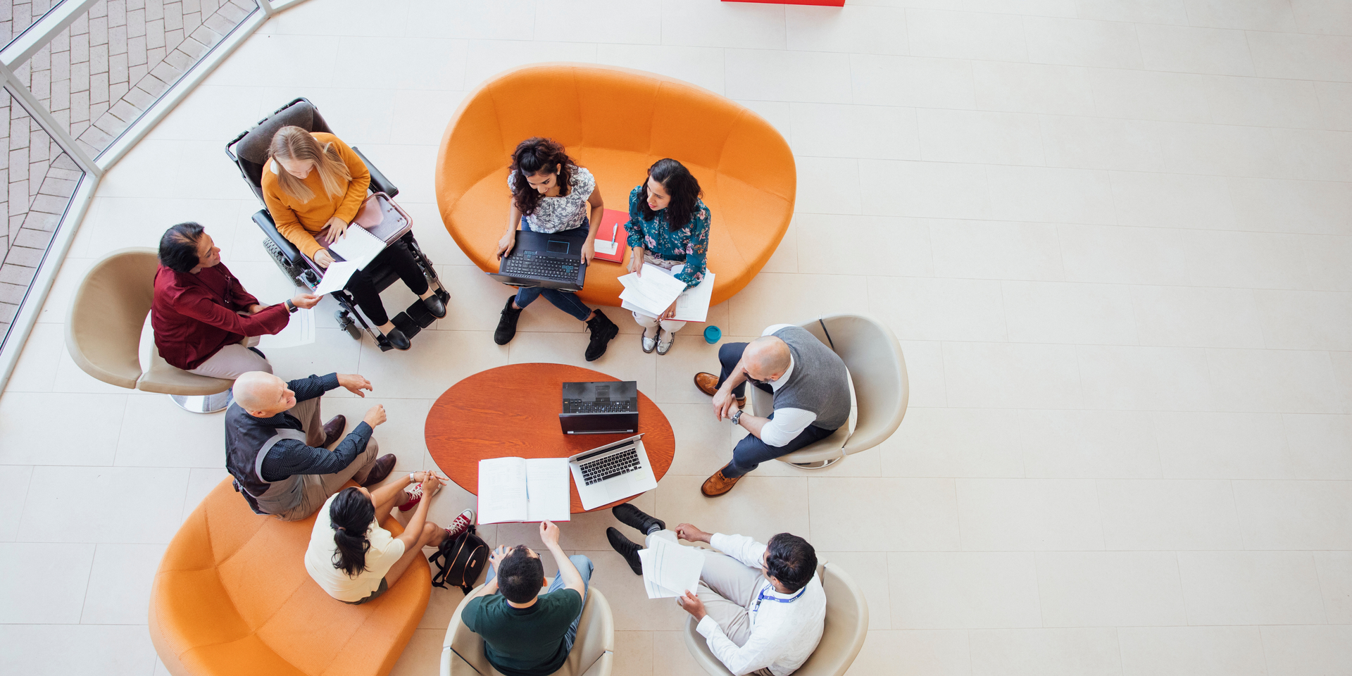 People in a circle, working and sitting on orange and tan chairs and a round table. Modern interior.