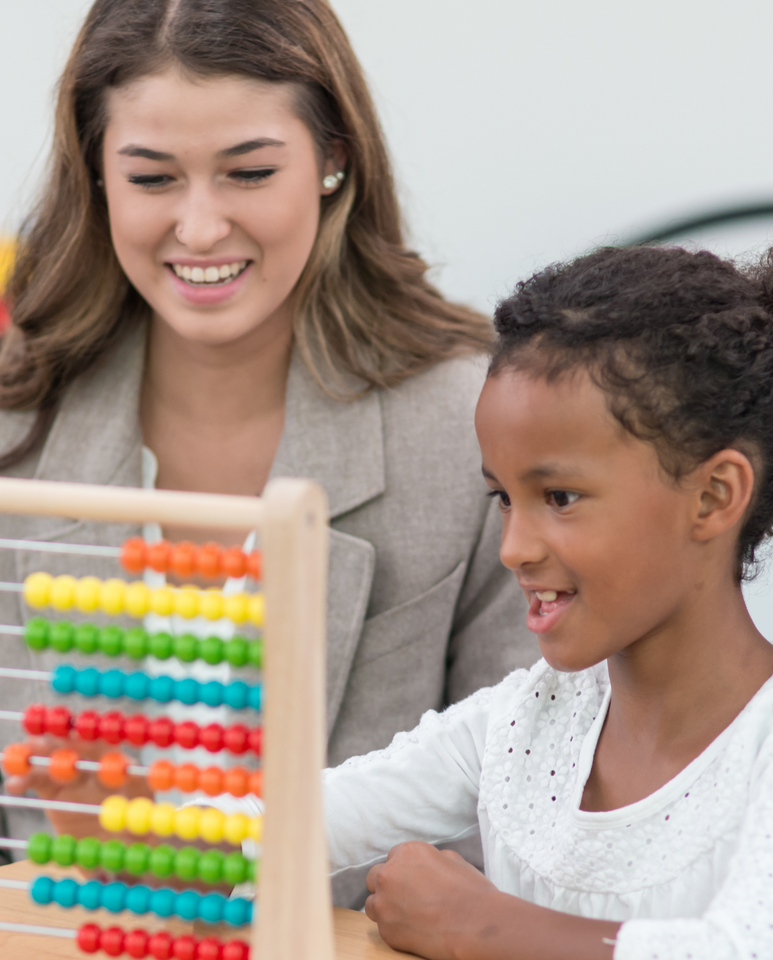 Woman in pink jacket assists smiling girl with schoolwork at a desk.