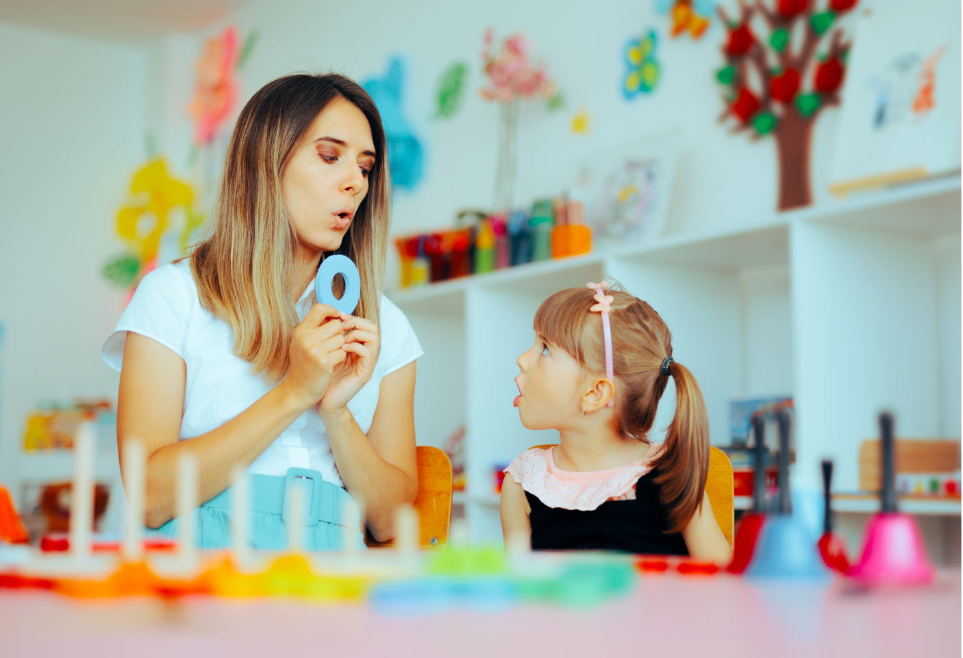 Woman demonstrates mouth shape with girl. Classroom setting with colorful toys and decorations.