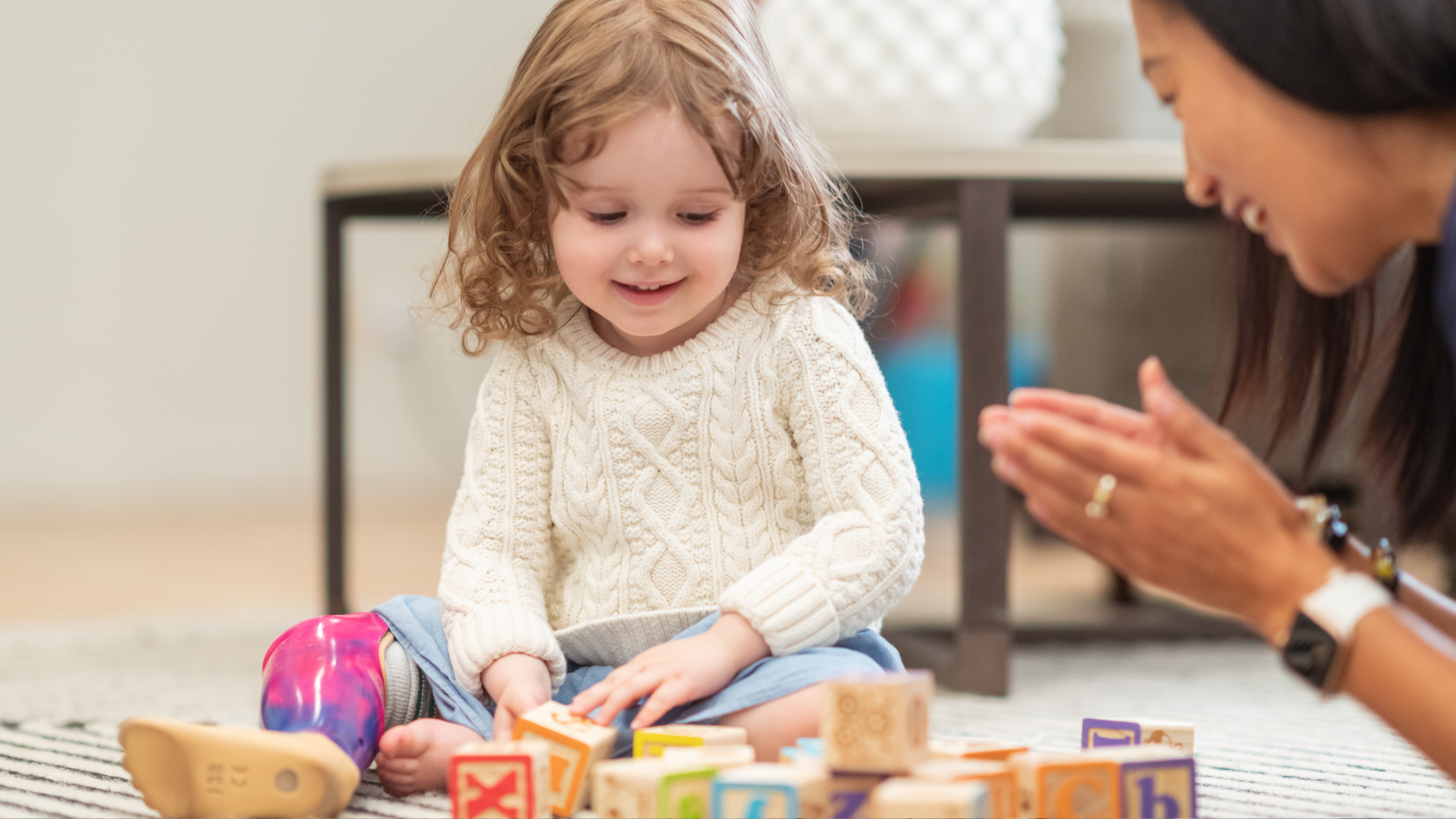 Child playing with blocks, smiling, while an adult claps nearby.