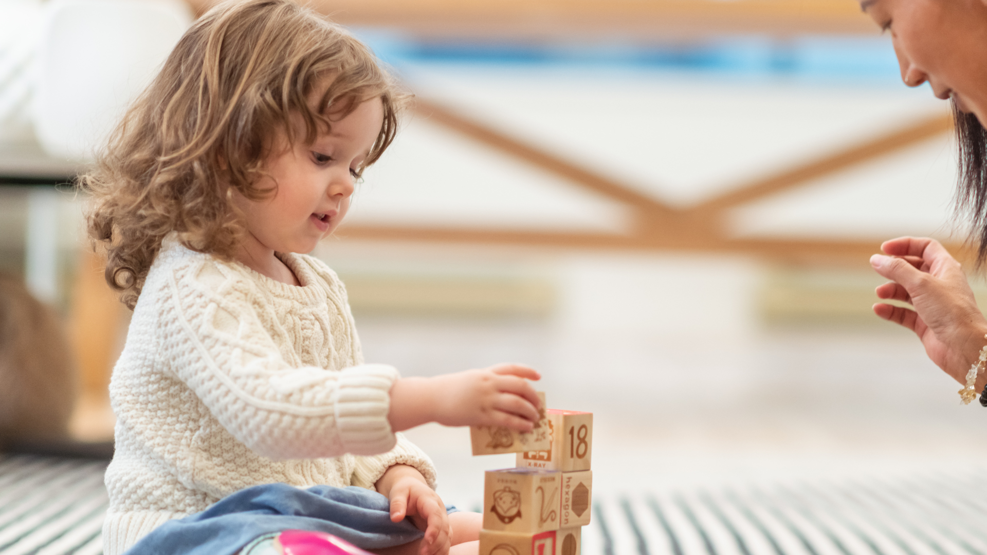 Child building tower of wooden blocks; person watches nearby.
