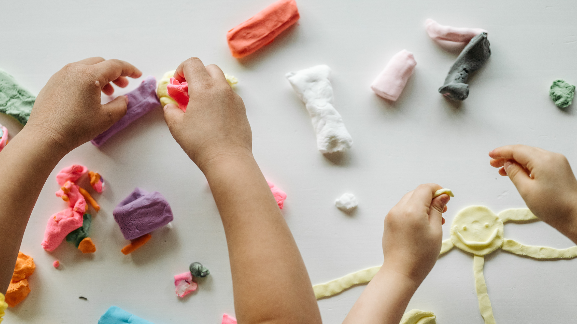 Hands of children playing with colorful clay on a white surface.