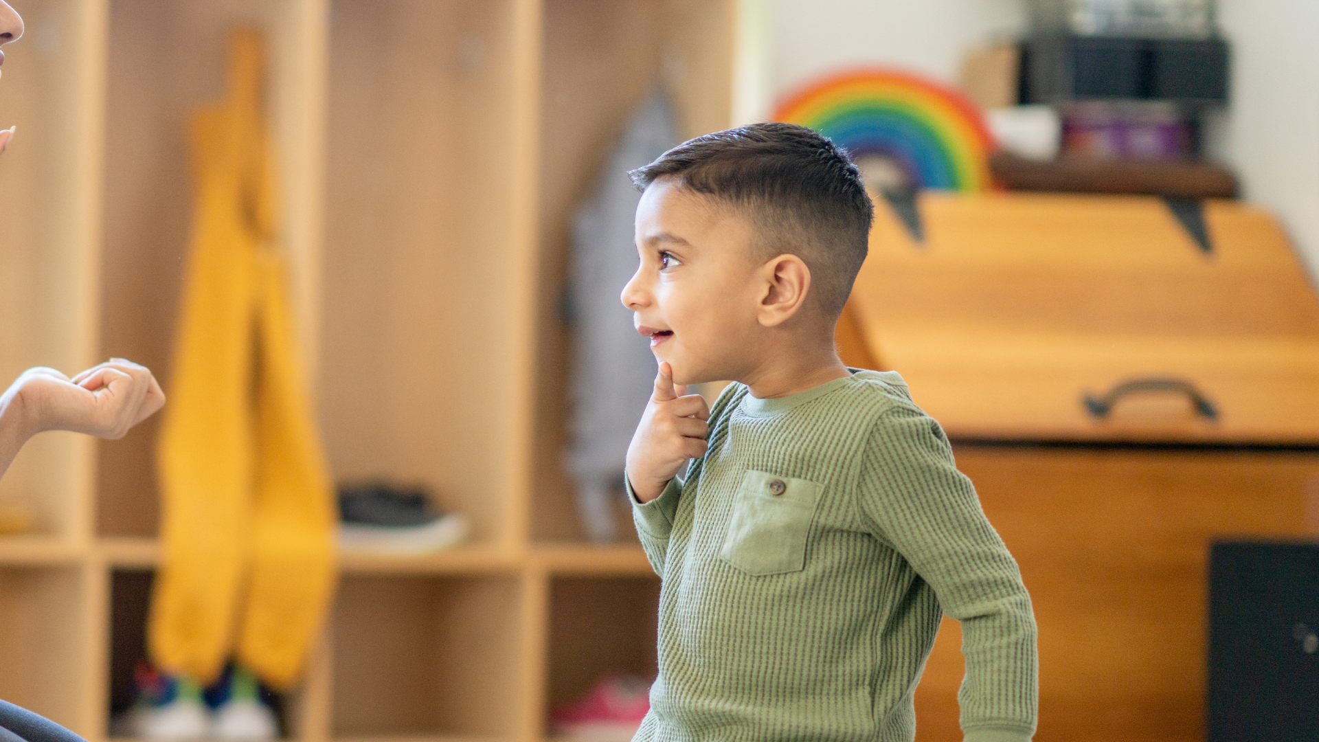 Boy in green shirt talking in a classroom, with wooden cubbies and rainbow in the background.