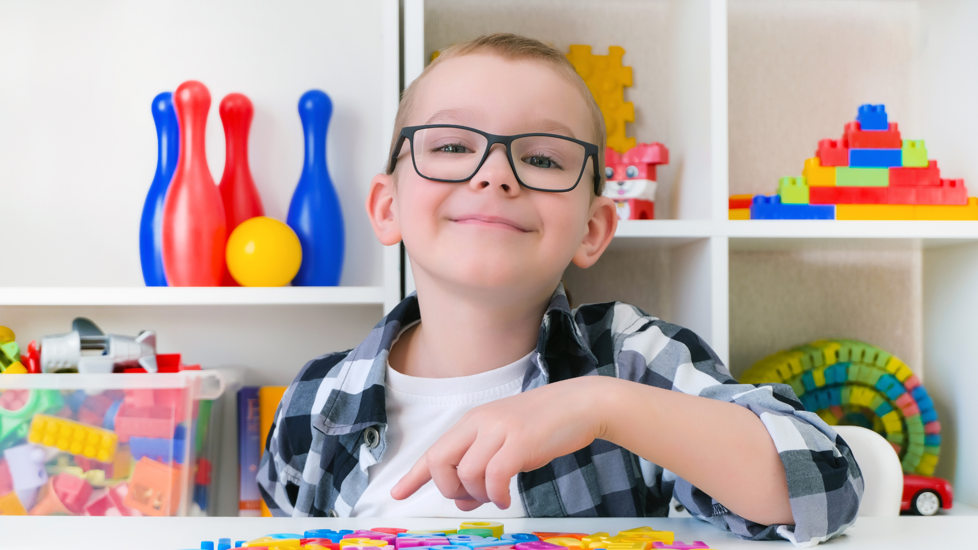 Boy in glasses smiles at the camera, surrounded by colorful toys.