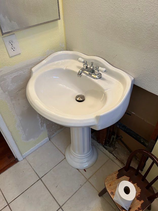 A white pedestal sink in a bathroom next to a chair and a roll of toilet paper.
