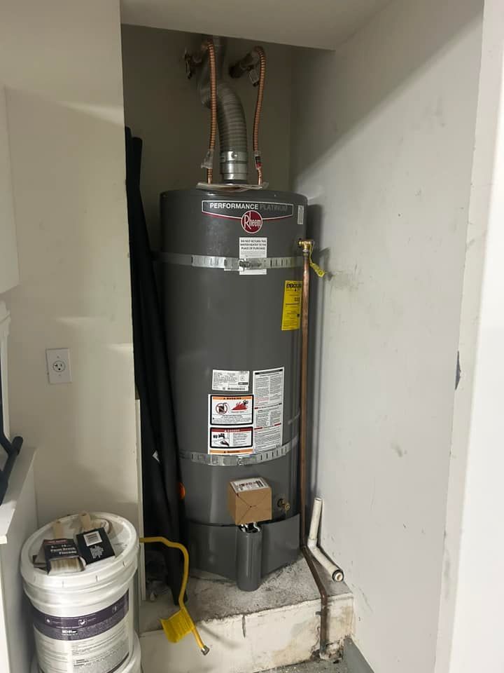 A water heater is sitting in a corner of a room next to a bucket of paint.