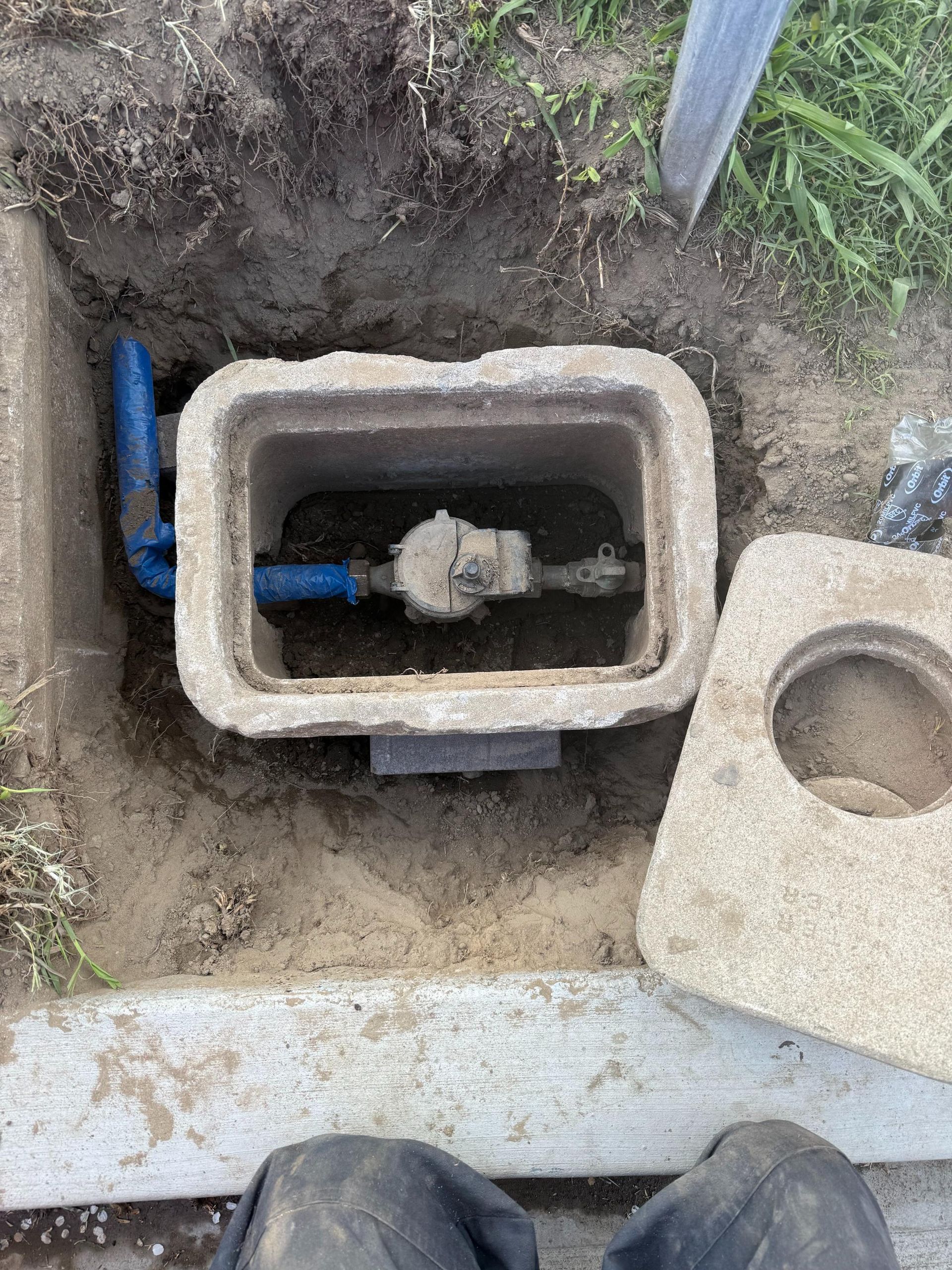 A person is standing next to a water meter in the dirt.