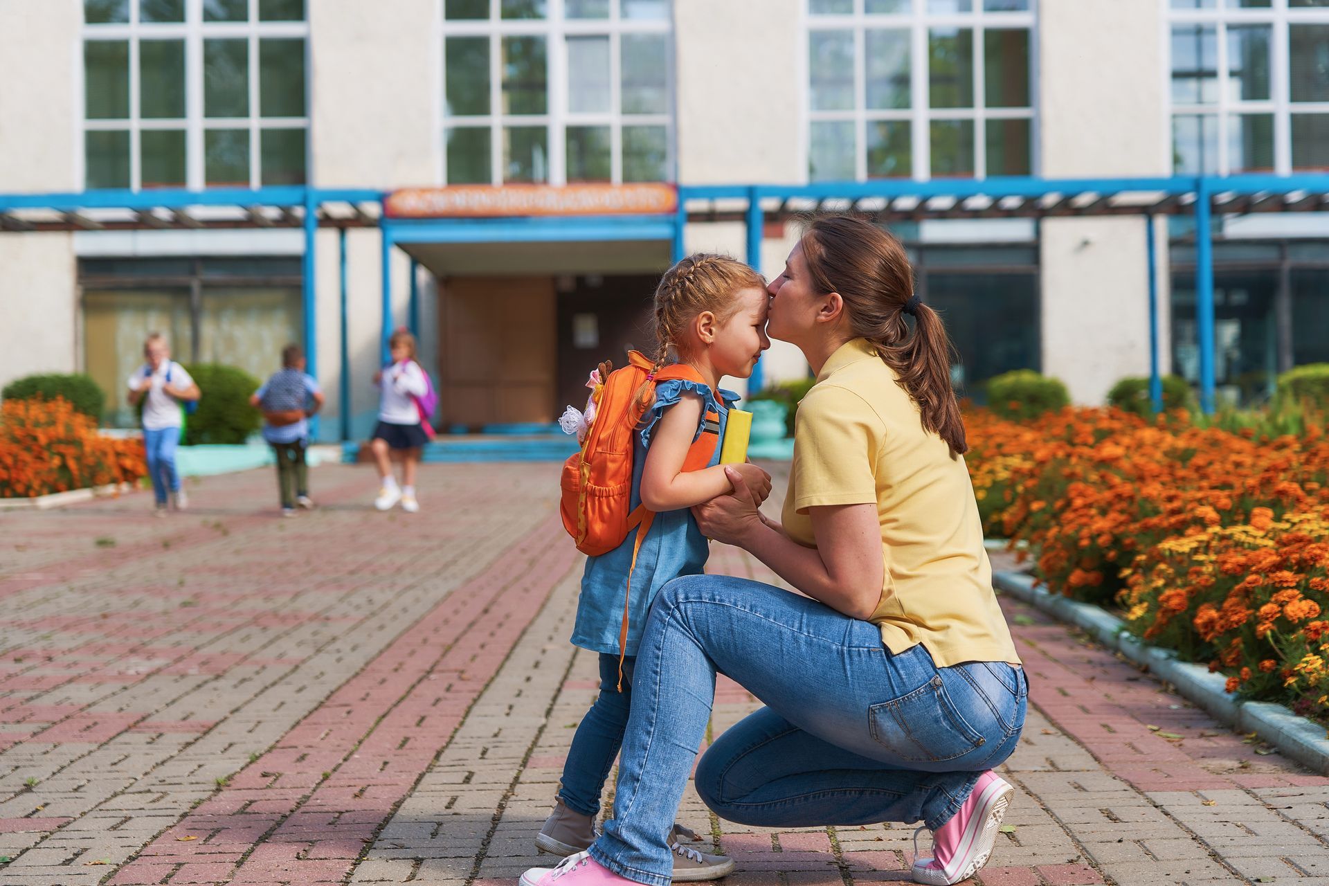 Woman kneels to kiss child's forehead outside a school. Child wears backpack, holds book. Flowers and children walking in background.