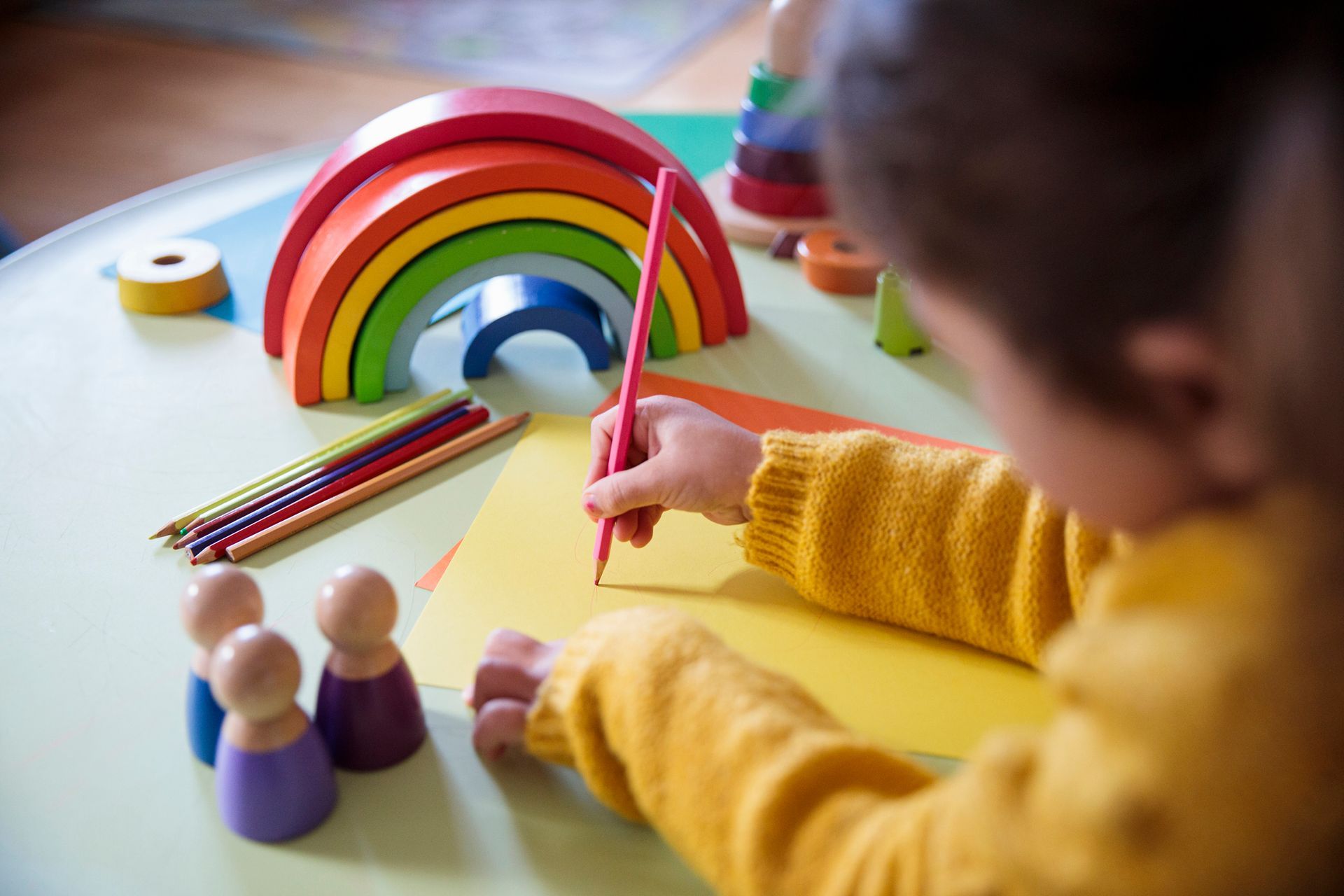 Child drawing with colored pencils beside wooden rainbow toys.