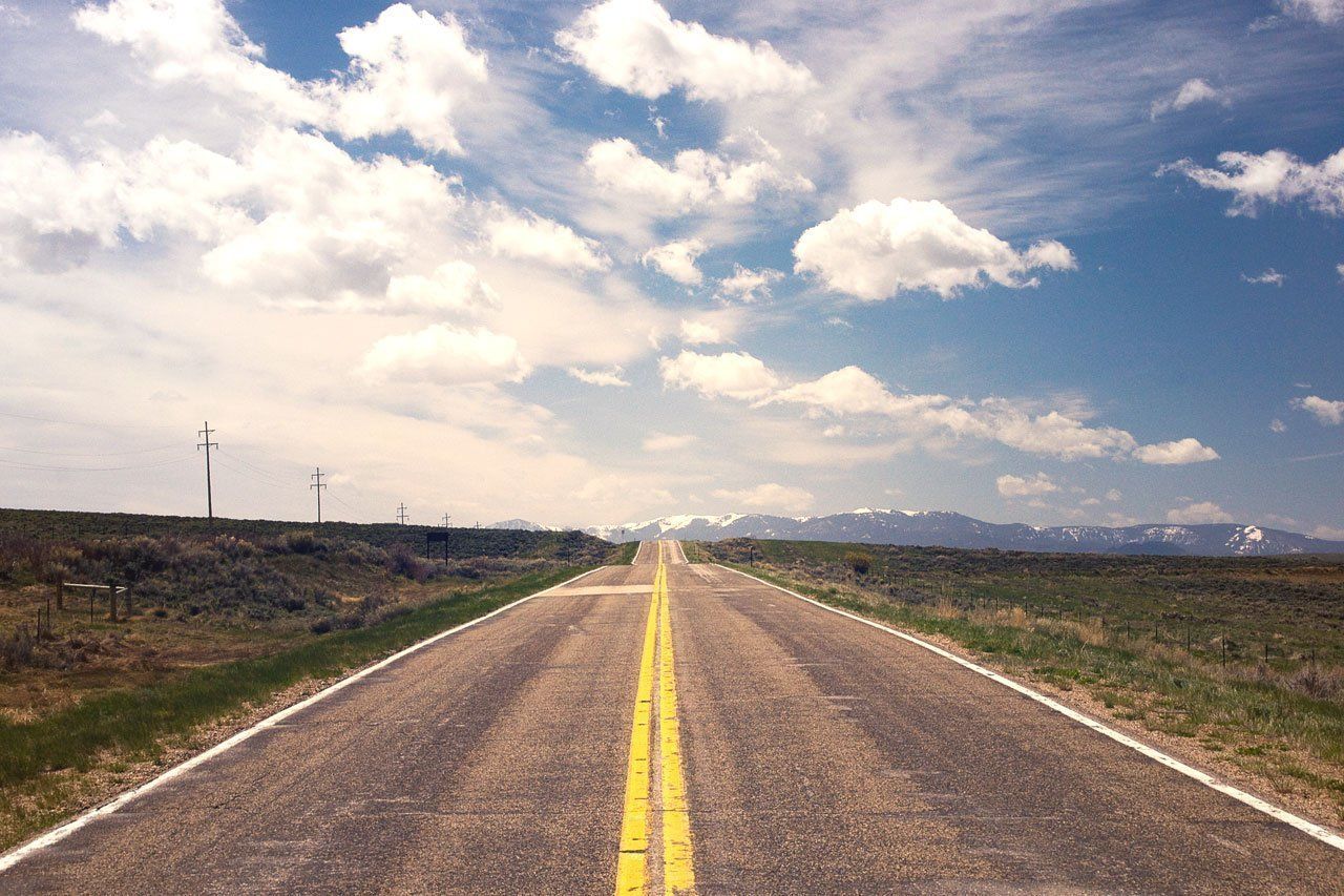 Road stretching towards distant mountains under a blue sky with fluffy white clouds.