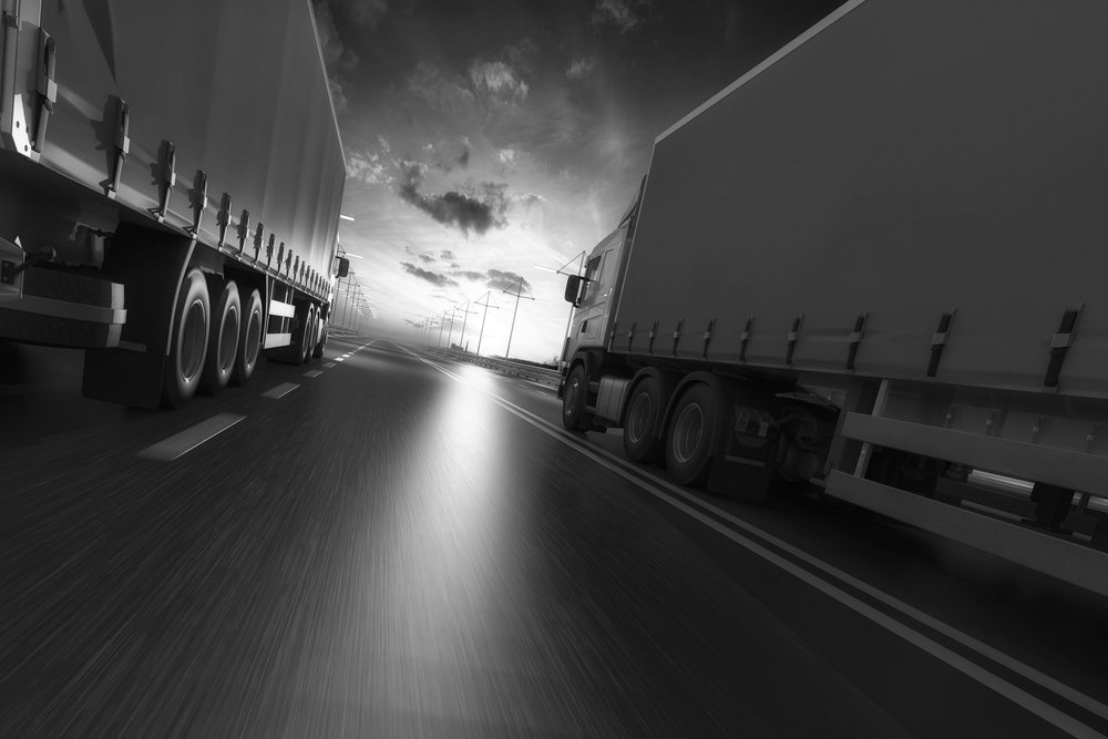 Two semi-trucks driving on a highway, dark sky with some wind turbines in the distance.