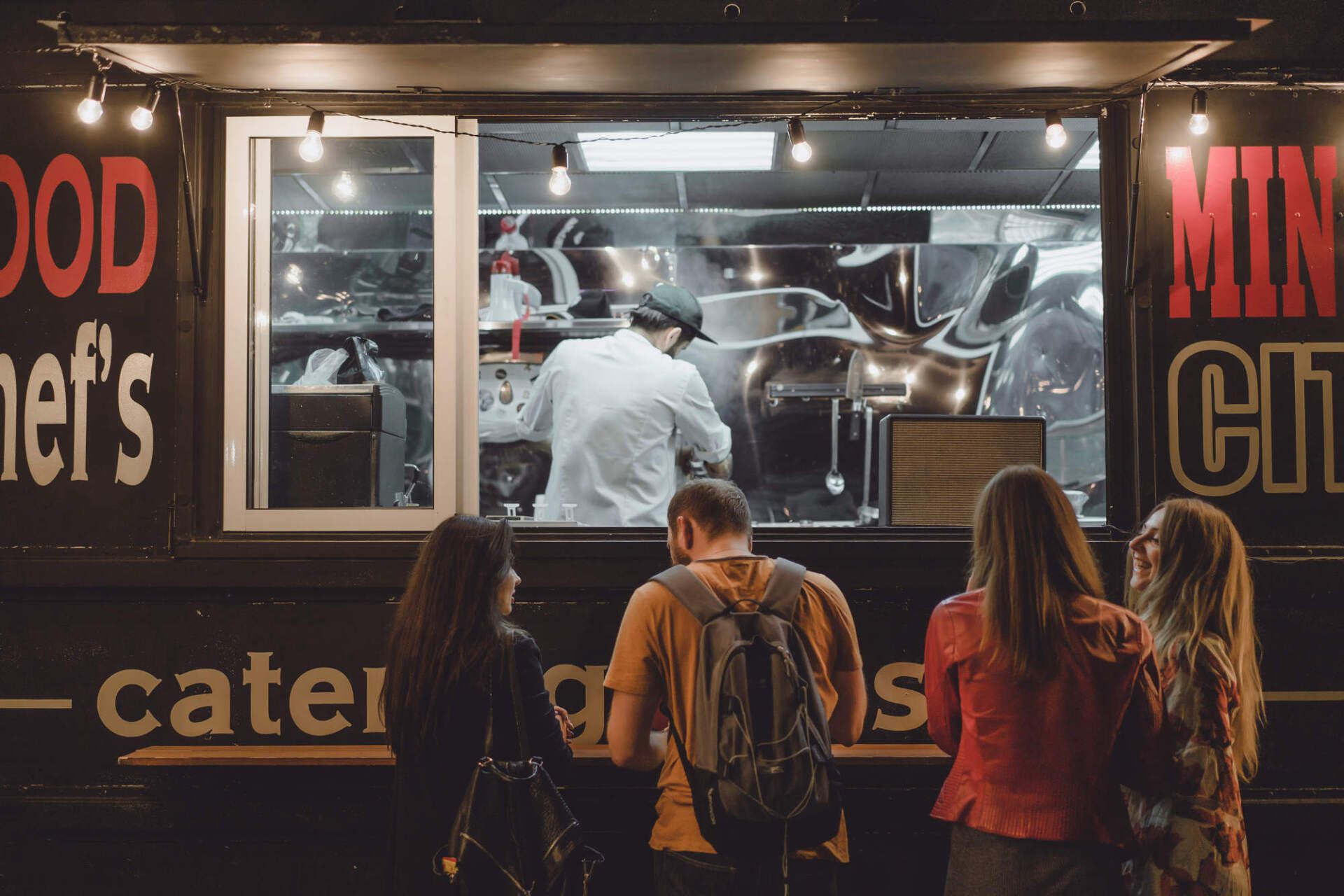 People ordering food from a food truck at night; chef works inside.