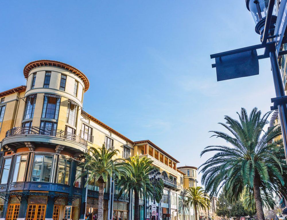 A sunny street with palm trees and ornate buildings, under a blue sky.