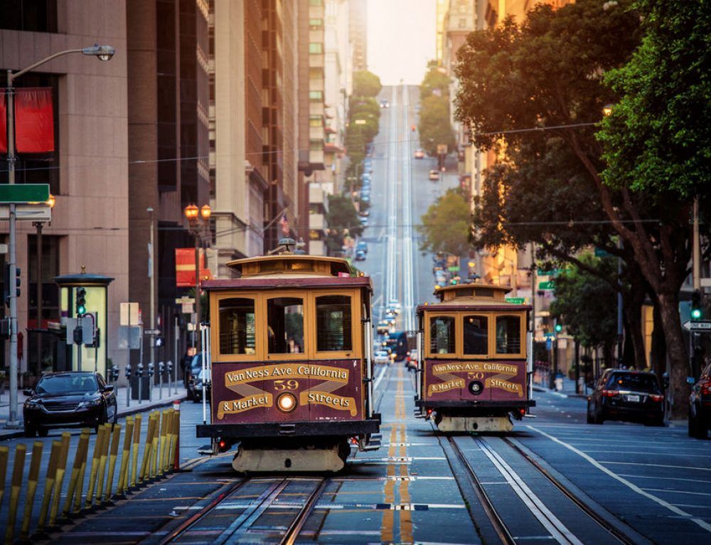 Two cable cars on a steep San Francisco street. Sunlight bathes the buildings.
