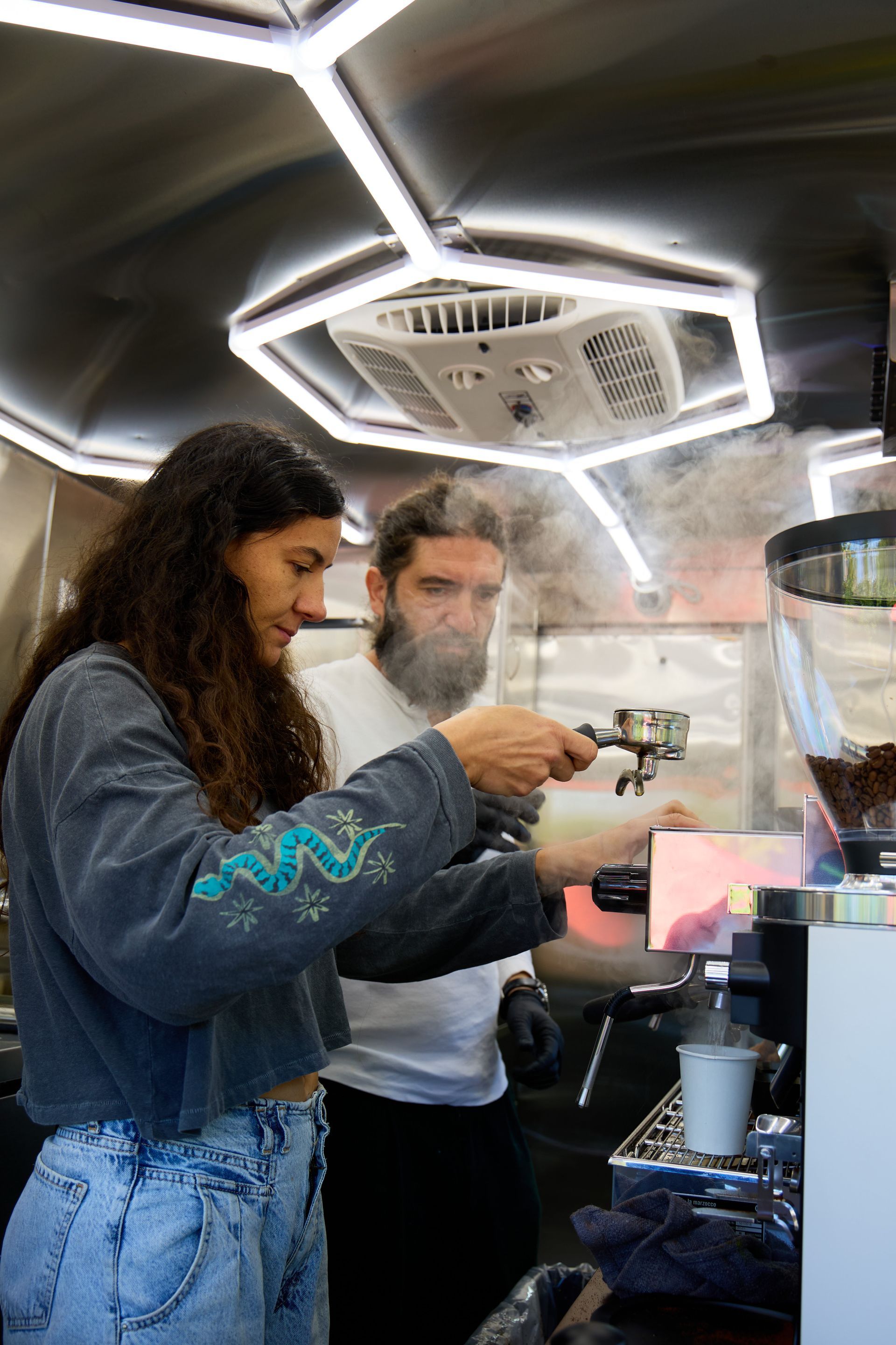 Two people making coffee in a food truck. Woman holding portafilter, man looking on. Steam visible.