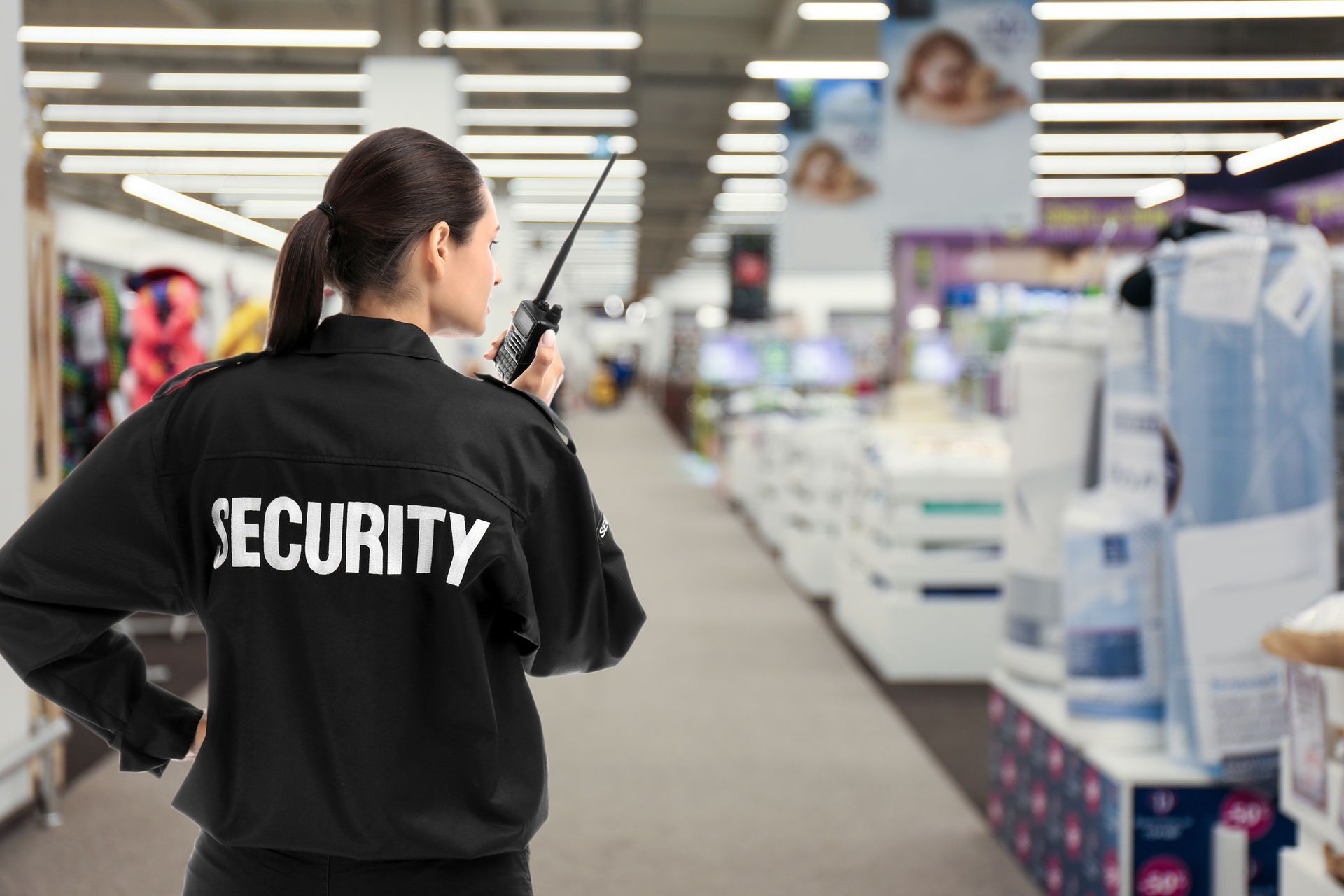 Security officer in black uniform using a walkie-talkie while patrolling a store aisle.