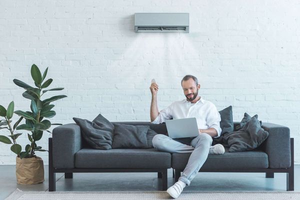 Handsome Man Turning On Air Conditioner With Remote Control While Using Laptop On Sofa At Home — Keizer, OR — Arrows Home Air