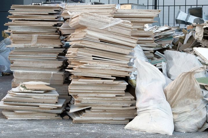 Two large stacks of discarded wooden particle board tiles and two white construction waste bags on a gravel surface.