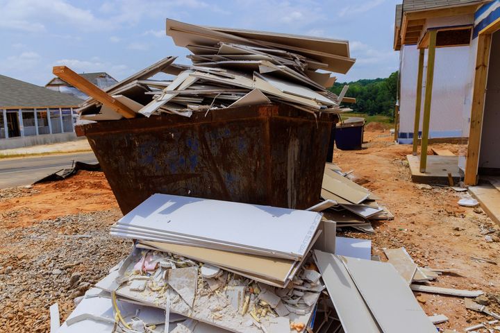 A metal dumpster overflowing with construction debris, including scraps and sheets of drywall, at a building site.