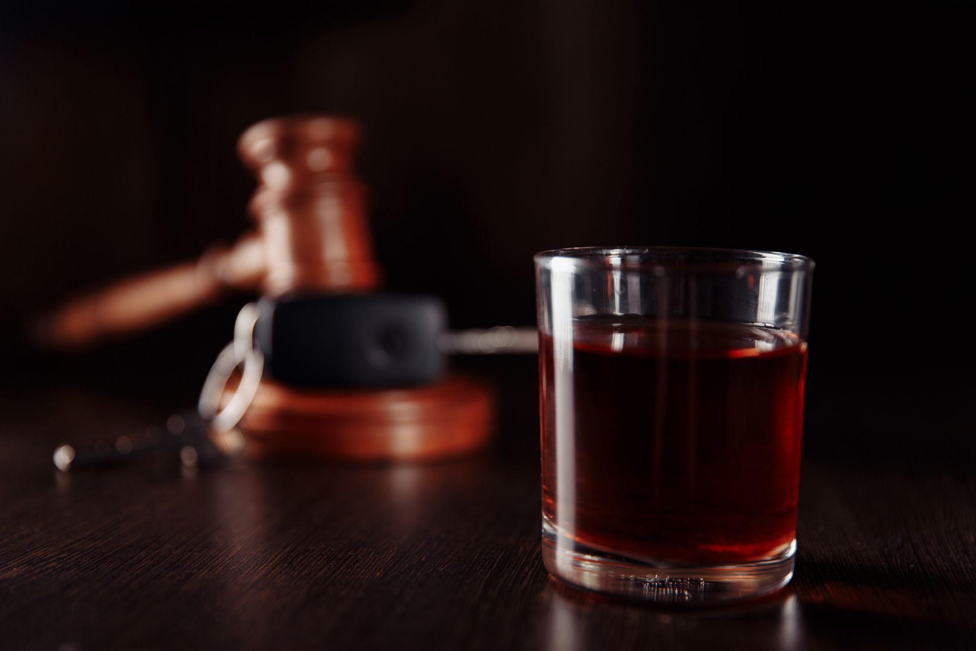 Shot glass of amber liquid, car keys, and gavel on a wooden surface, suggesting legal issues related to alcohol and driving.