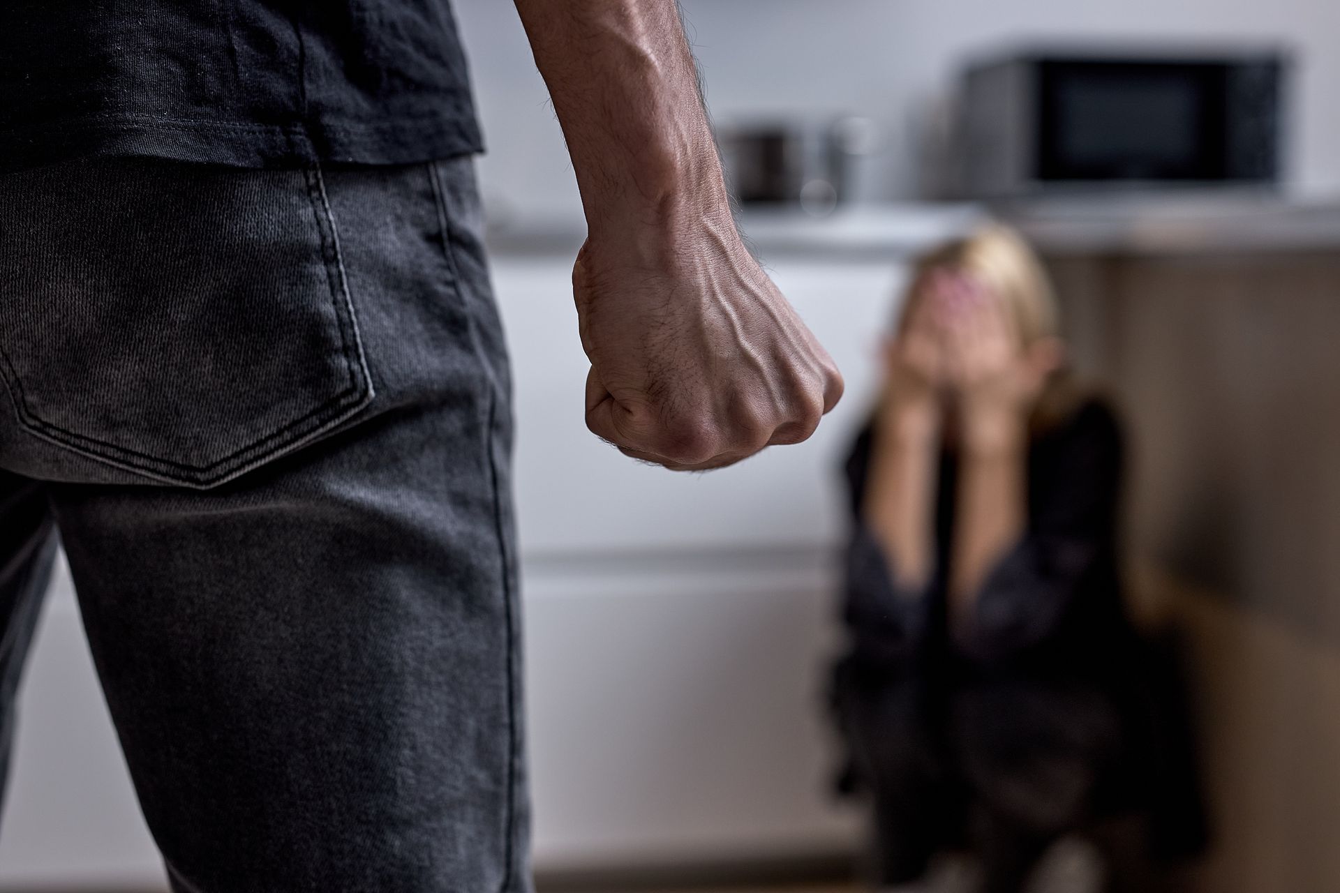 Man with fist raised, woman in corner covering face, kitchen setting.