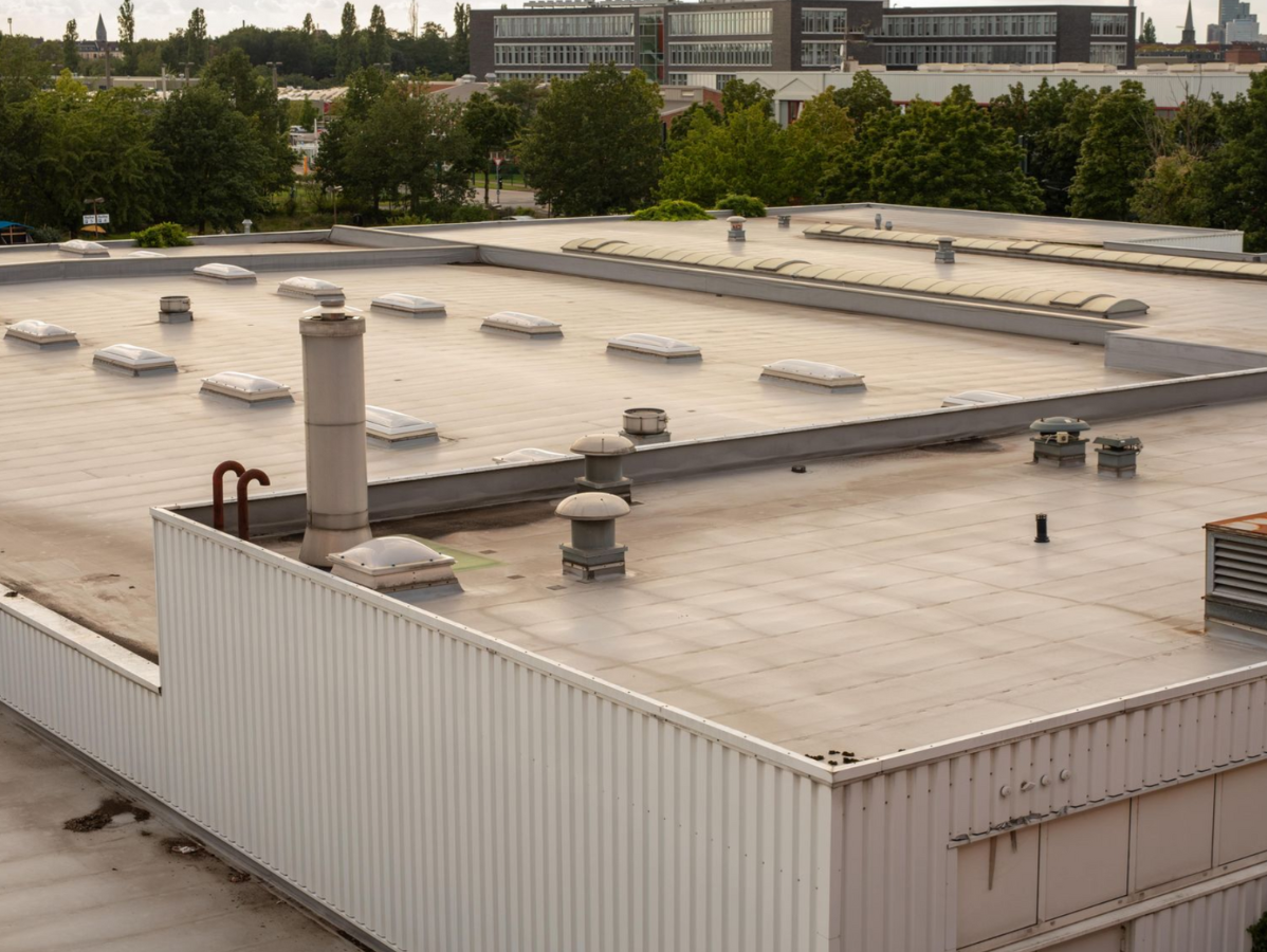 Flat commercial roof with vents, skylights, and a chimney. Buildings and trees in the background.