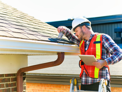 Construction worker in safety vest inspects a roof, using a tablet, on a sunny day.