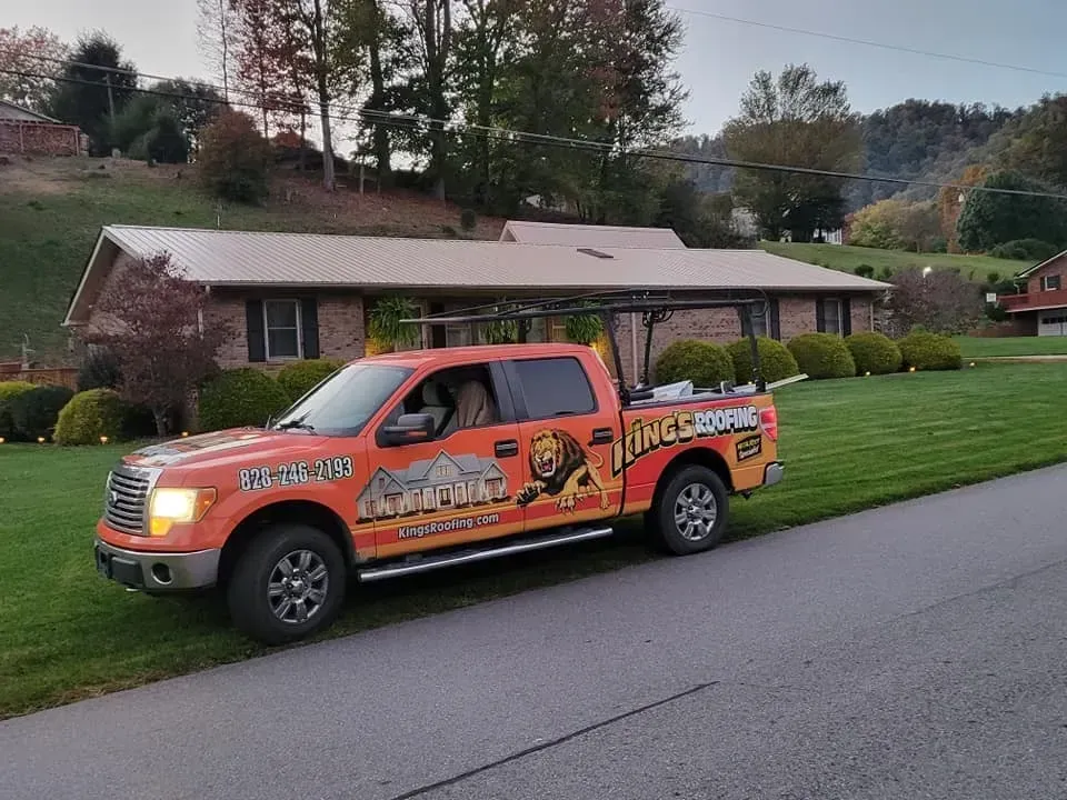 Orange work truck parked on a road in front of a house with a brown metal roof.
