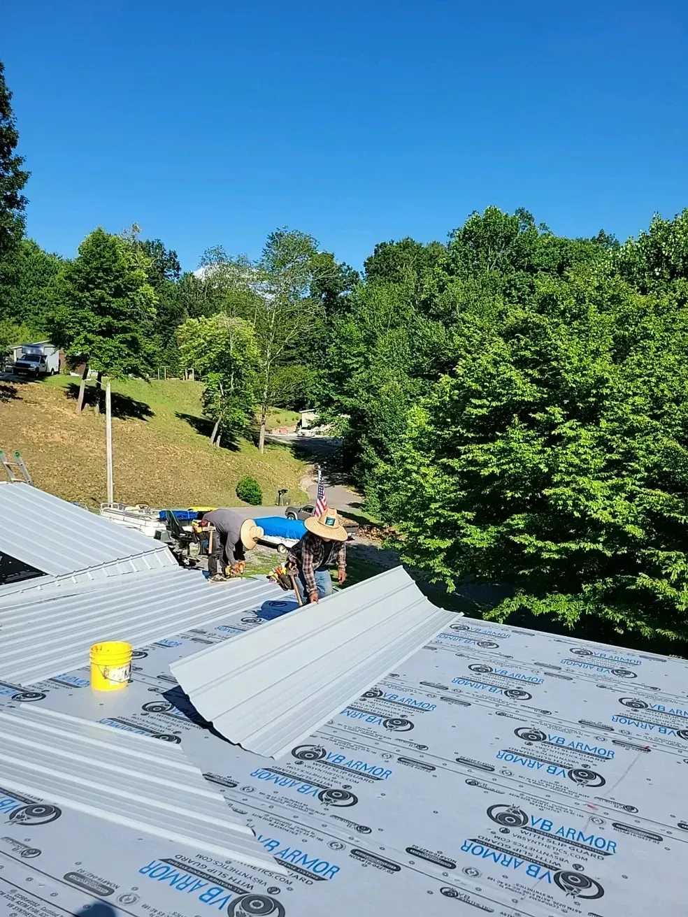 Roofers installing metal roofing on a sunny day with green trees in the background.