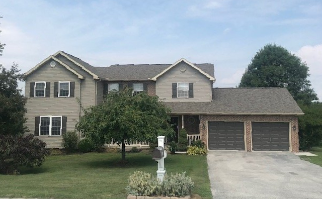 A two-story, tan-sided suburban home with a gray shingled roof, two-car garage, and a manicured front lawn.