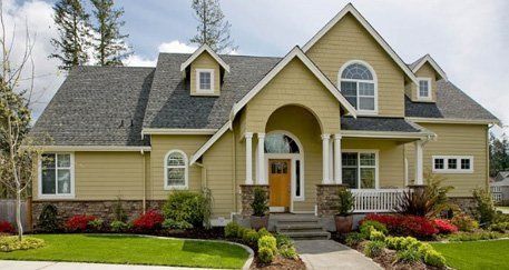 A two-story tan house with a gray roof, stone accents on the lower level, a front porch, and a manicured green lawn.