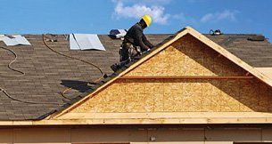 A construction worker in a yellow hard hat installs roofing shingles on a wooden-framed gable roof under a blue sky.