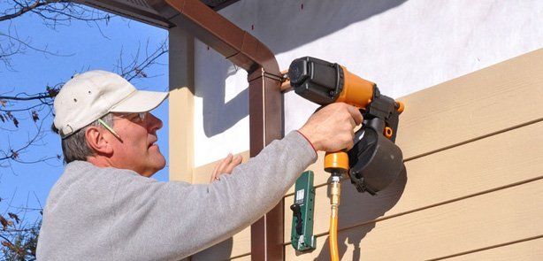 A person in a light-colored cap uses a nail gun to attach horizontal siding to an exterior wall.