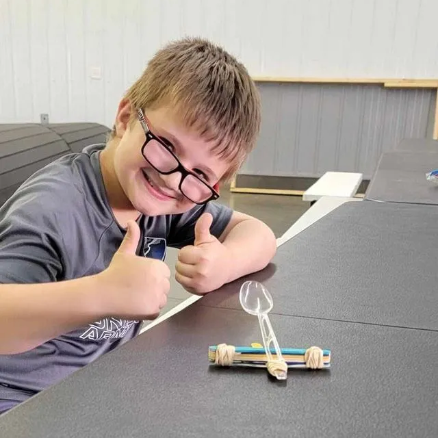 A young boy wearing glasses is giving a thumbs up while sitting at a table.
