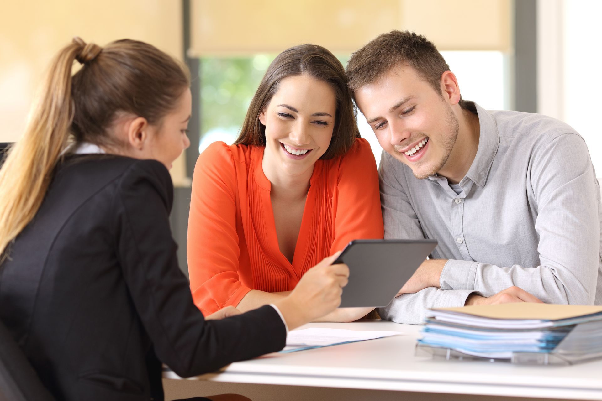 Three people looking at tablet. Smiling couple, woman in blazer, seated at a desk.