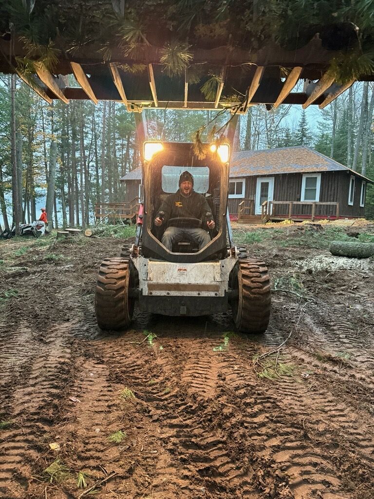 Bobcat skid steer with driver on a muddy path clearing brush near a cabin and lake.