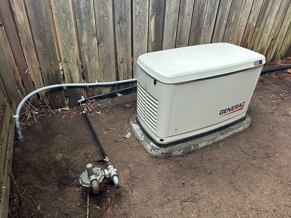 A generac generator is sitting on the ground next to a wooden fence showing the wiring and gas line pipe.