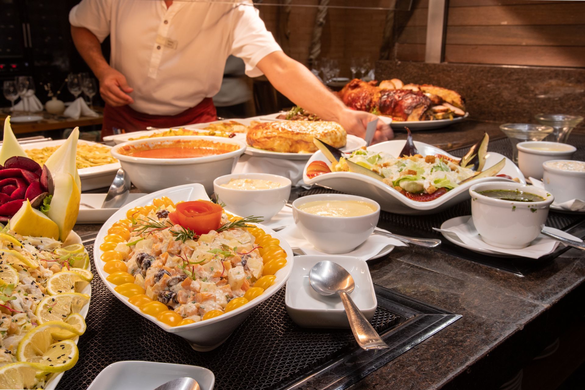 Uma mesa de bufê com tigelas de saladas, acompanhamentos e um assado como prato principal, com uma pessoa servindo a comida.