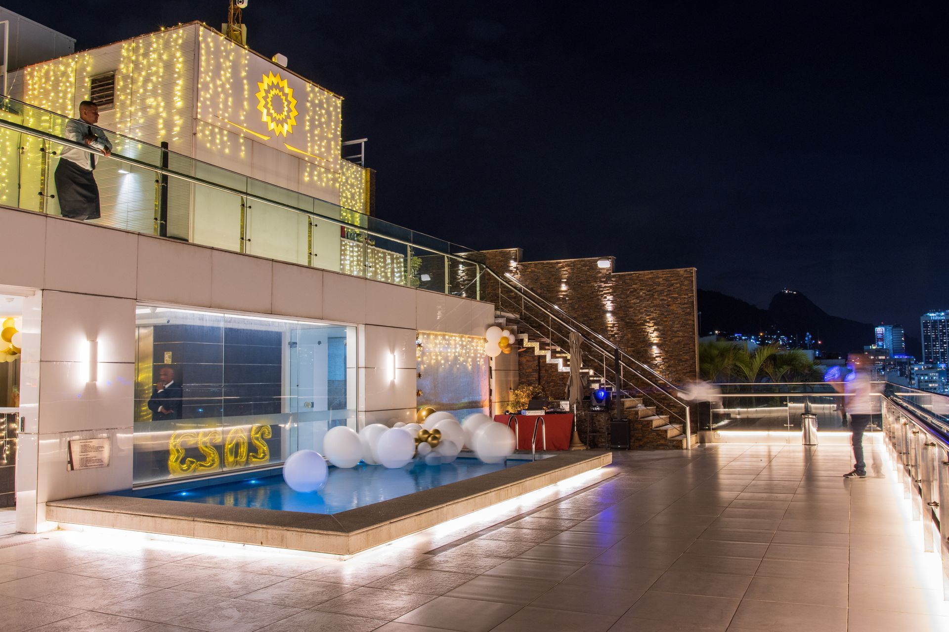 Vista noturna de um deck externo em níveis com piscina iluminada, arquitetura moderna e luzes de corda em um navio de cruzeiro.