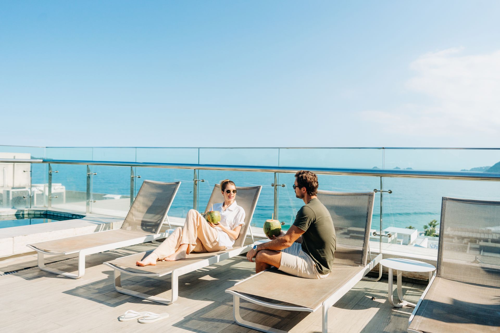 Um casal relaxa em espreguiçadeiras num terraço ensolarado de um resort com vista para o oceano, enquanto seguram bebidas.