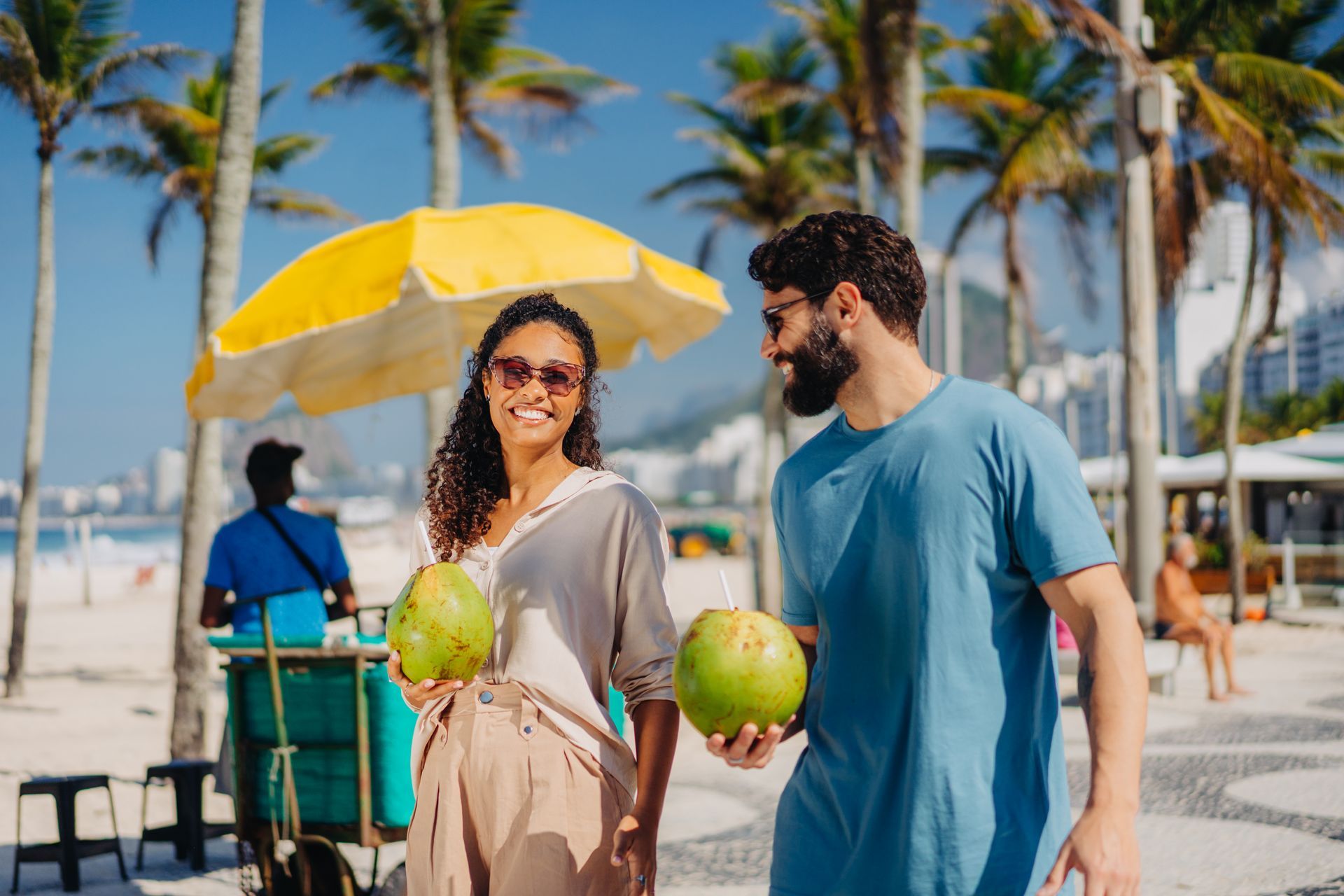 Um casal caminha por um calçadão tropical iluminado pelo sol, sorrindo e segurando cocos verdes sob um guarda-chuva amarelo brilhante.