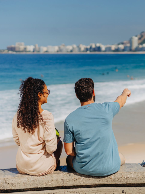 Duas pessoas sentam-se na praia, olhando para o oceano e apontando para a costa distante.