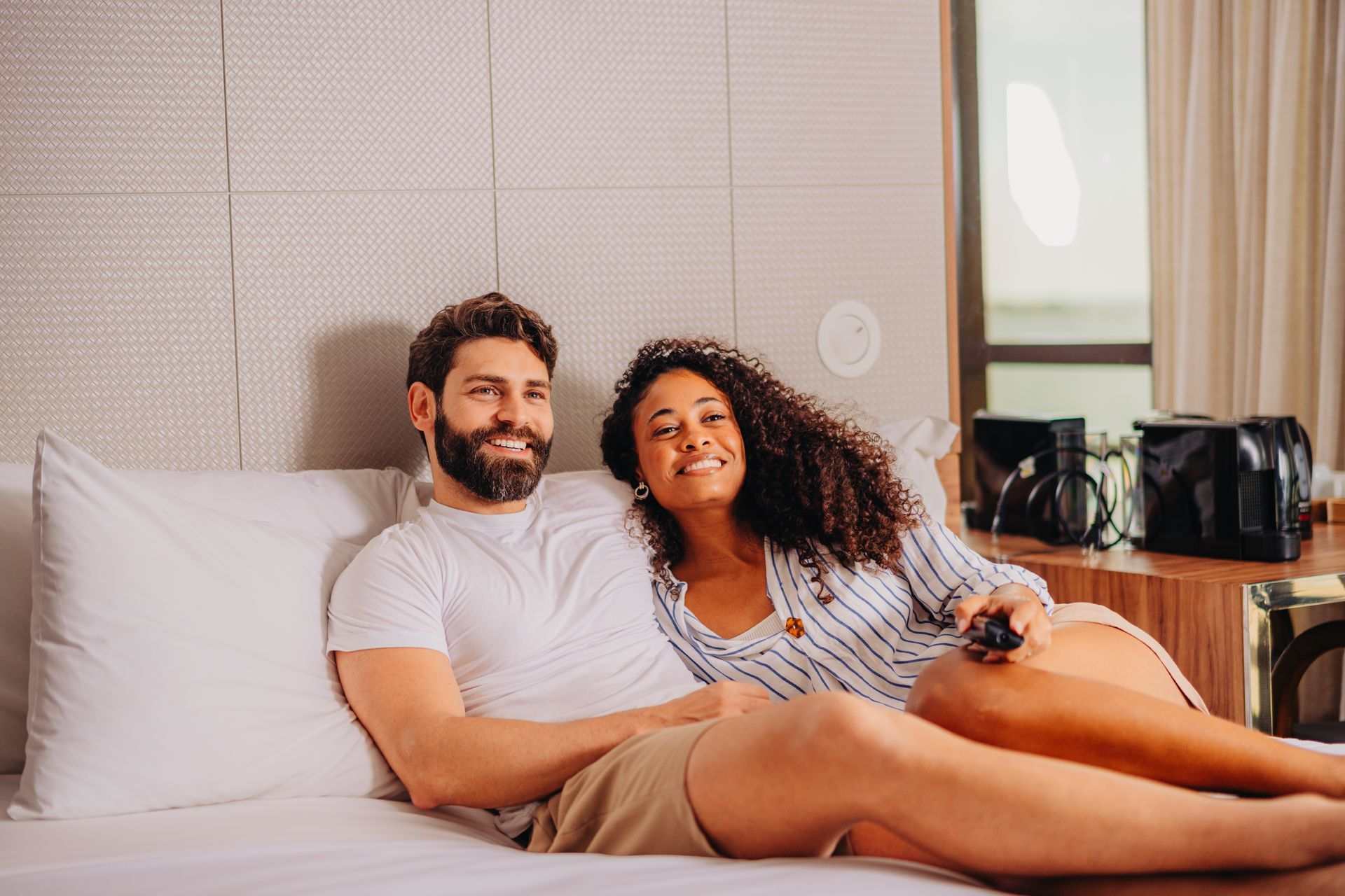 Um casal sorri enquanto relaxa em uma cama, segurando um controle remoto e assistindo televisão em um quarto de hotel moderno.