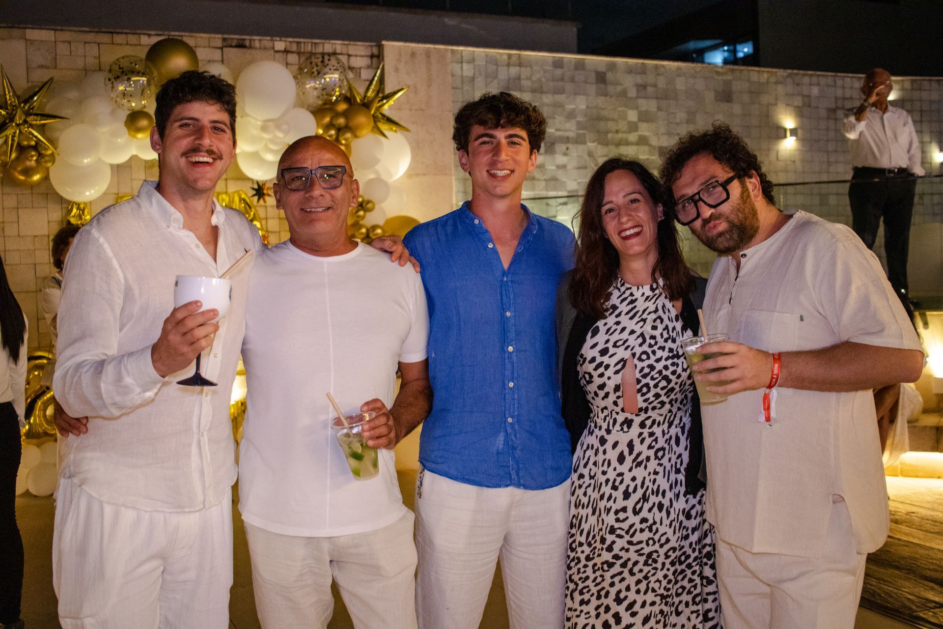 Cinco pessoas sorriem em uma festa ao ar livre com decoração branca, segurando bebidas enquanto posam para uma foto em grupo.