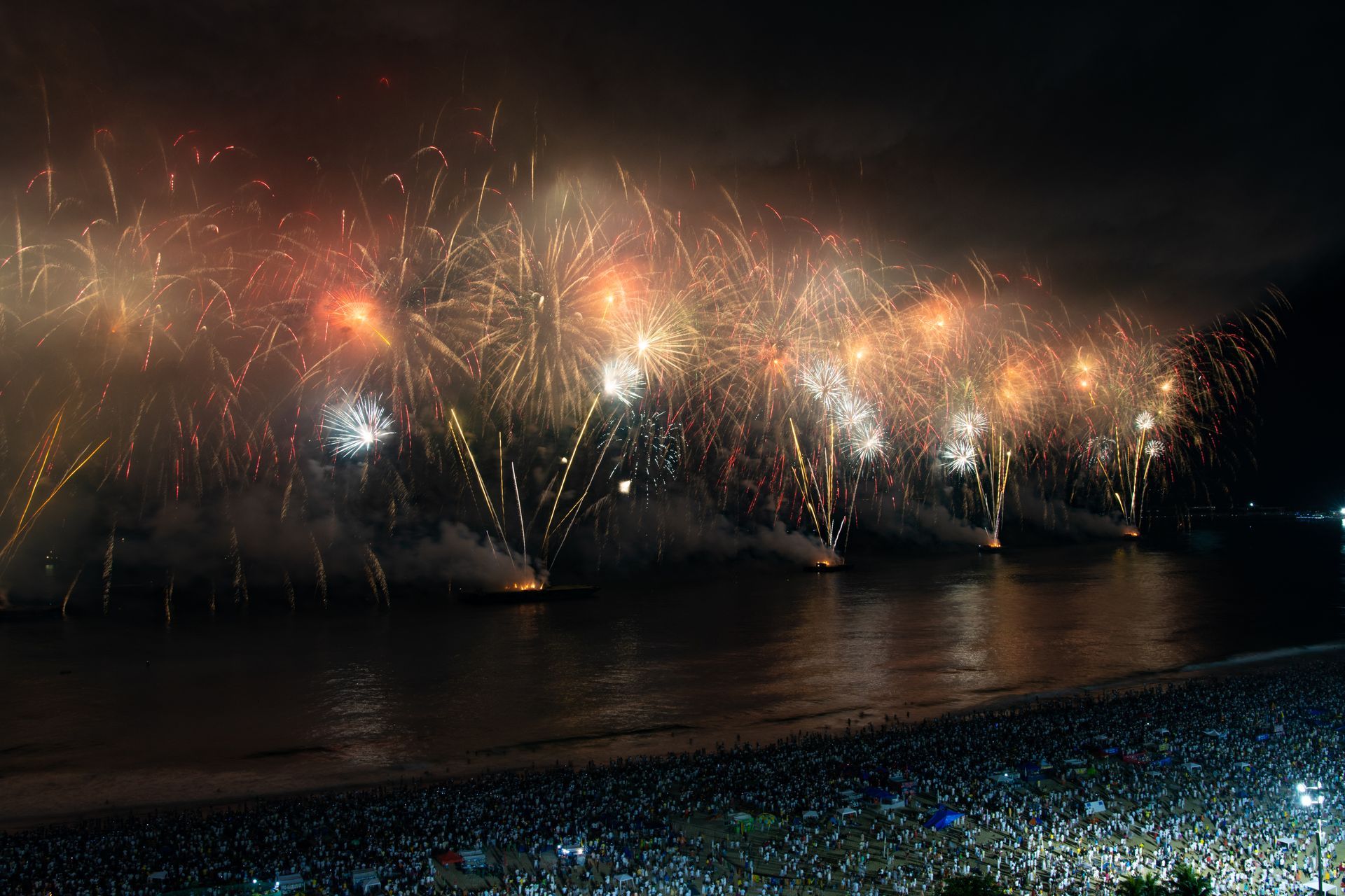Um grande espetáculo de fogos de artifício ilumina o céu noturno sobre uma praia lotada de espectadores.